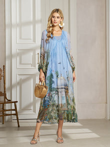 A woman stands indoors wearing the Social Queen Garden-Inspired Print Pleated Dress with long sleeves and a scenic design, accessorized with a small wicker bag, sandals, and large earrings.