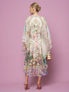 A woman faces away in the Garden Print Silk Dress by Social Queen—a flowy, pastel floral silk dress with a waist tie—paired with pink heels and a gold handbag, set against a pink background.