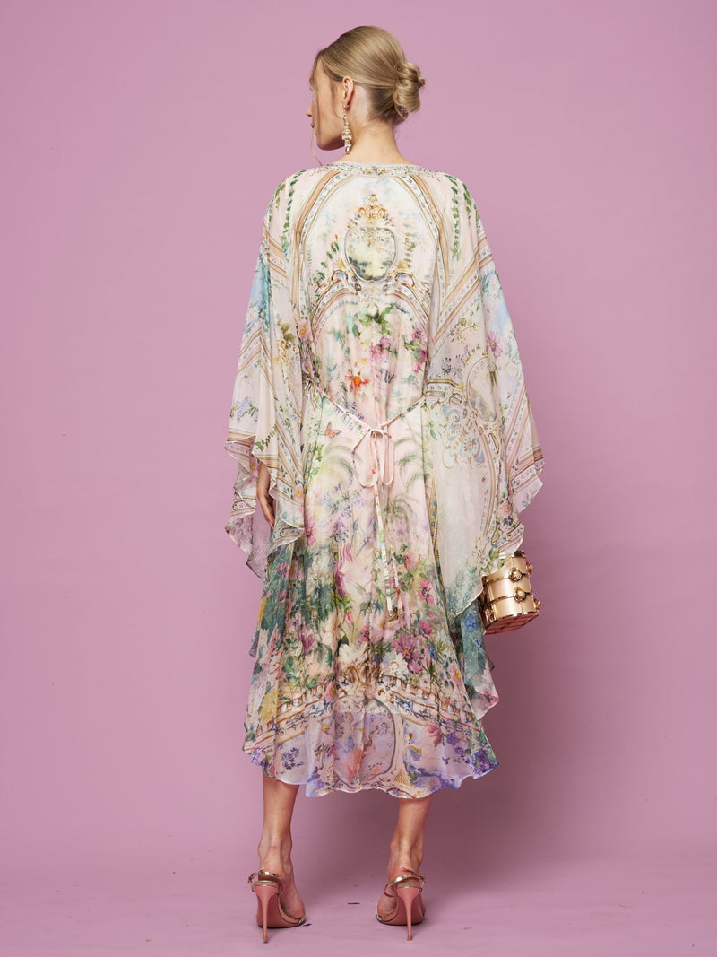 A woman faces away in the Garden Print Silk Dress by Social Queen—a flowy, pastel floral silk dress with a waist tie—paired with pink heels and a gold handbag, set against a pink background.