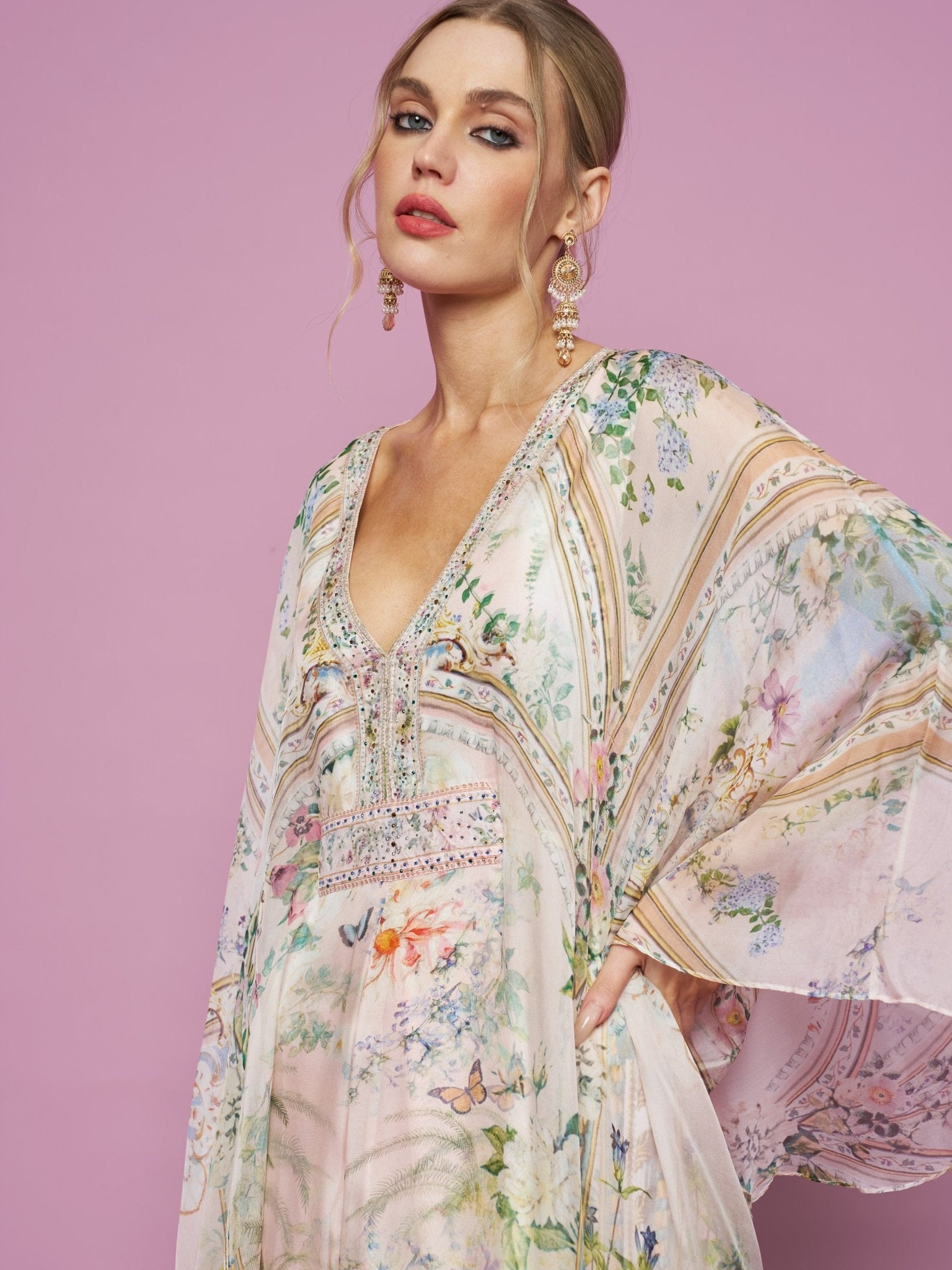 Model in Social Queen's Garden Print Silk Dress, featuring a lightweight silk floral design and deep V-neck, poses before a pink backdrop with chandelier earrings, gazing at the camera.
