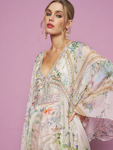 Model in Social Queen's Garden Print Silk Dress, featuring a lightweight silk floral design and deep V-neck, poses before a pink backdrop with chandelier earrings, gazing at the camera.