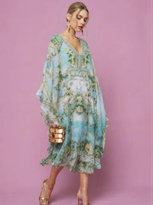 A woman in the Social Queen Garden Print Silk Dress stands against a pink background, pairing the flowing blue and green floral silk with gold high heels and a matching gold clutch.