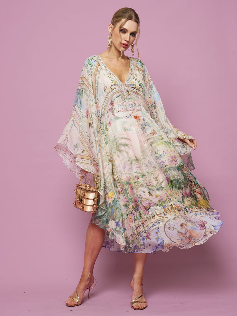 A woman stands against a pink background in the Garden Print Silk Dress by Social Queen, featuring flowing lightweight silk and wide sleeves, styled with a metallic purse and strappy heels.