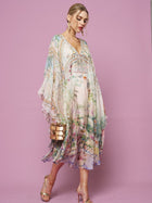 A woman in the Social Queen Garden Print Silk Dress, featuring a flowy pastel floral design and deep V-neck, stands against a light purple background, accessorized with a gold clutch and strappy heels.