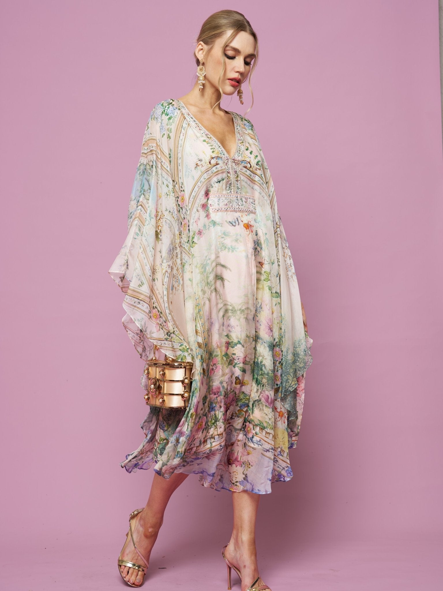 A woman in the Social Queen Garden Print Silk Dress, featuring a flowy pastel floral design and deep V-neck, stands against a light purple background, accessorized with a gold clutch and strappy heels.