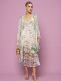 A woman in the Social Queen Garden Print Silk Dress, featuring flowy silk with floral and scenic designs, holds a small handbag while standing against a plain pink background.