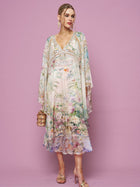 A woman in the Social Queen Garden Print Silk Dress, featuring flowy silk with floral and scenic designs, holds a small handbag while standing against a plain pink background.
