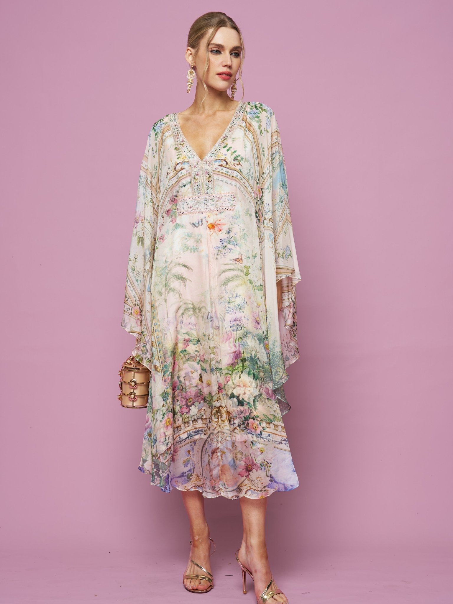 A woman in the Social Queen Garden Print Silk Dress, featuring flowy silk with floral and scenic designs, holds a small handbag while standing against a plain pink background.
