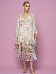 A woman in the Social Queen Garden Print Silk Dress, featuring flowy silk with floral and scenic designs, holds a small handbag while standing against a plain pink background.