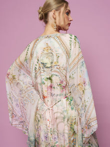 A woman stands against a pink background wearing the Garden Print Silk Dress by Social Queen, featuring wide sleeves, lightweight silk fabric, and a delicate floral print. Her hair is styled in a bun.