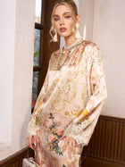 A woman stands indoors near a window wearing the Social Queen Garden Print Silk Dress with lace trim—an elegant choice for wedding guests—styled with statement earrings and a necklace.