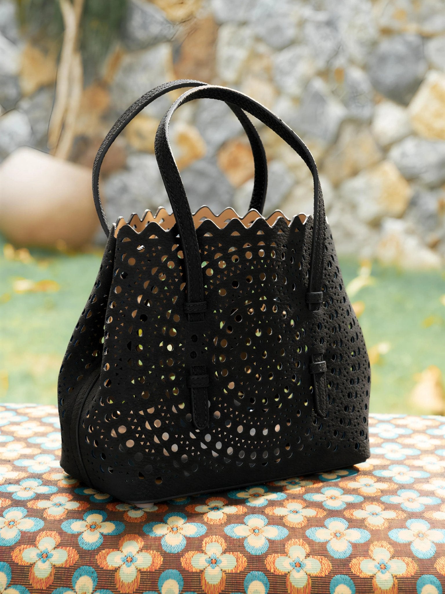 The Social Queen Geometric Cut-Out Leather Bag, featuring black genuine leather, a suede finish, scalloped edges, and cutout details, rests on a patterned outdoor surface with grass and a stone wall behind it.