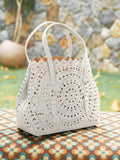 The Social Queen Geometric Cut-Out Leather Bag, in white genuine leather, is displayed outdoors on a patterned surface with a stone wall and grass behind it.