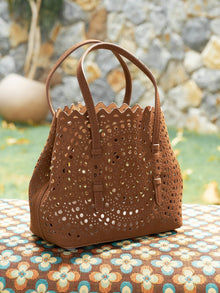 The Social Queen Geometric Cut-Out Leather Bag with dual handles is displayed outdoors on a patterned surface, set against a stone wall and greenery backdrop.
