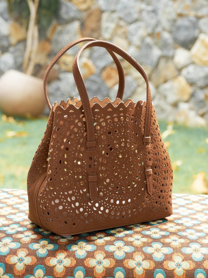 The Social Queen Geometric Cut-Out Leather Bag with dual handles is displayed outdoors on a patterned surface, set against a stone wall and greenery backdrop.