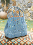 The Social Queen Geometric Cut-Out Leather Bag, in light blue with two handles, is displayed outdoors on a patterned surface, against a backdrop of a stone wall and greenery.