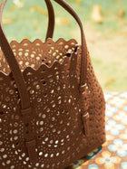 The Social Queen Geometric Cut-Out Leather Bag, featuring brown leather with geometric cutouts and scalloped edges, rests outdoors on a patterned surface among greenery and fallen leaves.