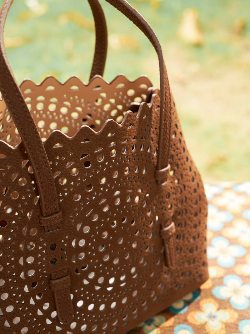 The Social Queen Geometric Cut-Out Leather Bag, featuring brown leather with geometric cutouts and scalloped edges, rests outdoors on a patterned surface among greenery and fallen leaves.