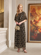 A woman wears the Social Queen Gilded Beaded Cape Dress indoors, holding a small handbag with a large framed painting behind her—an elegant choice for formal cocktail events.