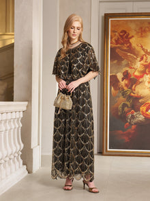 A woman wears the Social Queen Gilded Beaded Cape Dress indoors, holding a small handbag with a large framed painting behind her—an elegant choice for formal cocktail events.