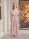 A woman stands indoors near a railing and a large angel painting, wearing the Social Queen Gilded Beaded Cape Dress in light pink.