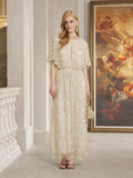 A woman stands indoors near a classical painting, wearing the Social Queen Gilded Beaded Cape Dress—a floor-length, light-colored formal gown—while holding a matching small bag.