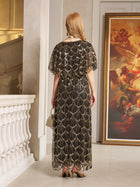 A blonde woman stands indoors facing away, wearing the Social Queen Gilded Beaded Cape Dress and holding a small handbag. A large framed painting is visible on the right.