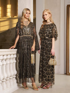 Two women wear Social Queen's Gilded Beaded Cape Dress indoors, each holding a small handbag and wearing gold heels—a chic choice for an elegant evening or formal cocktail event.