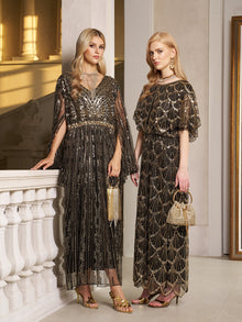 Two women wear Social Queen's Gilded Beaded Cape Dress indoors, each holding a small handbag and wearing gold heels—a chic choice for an elegant evening or formal cocktail event.