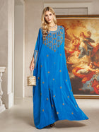 A woman wears the Gilded Leaf Embellished Cape Dress by Social Queen, a royal blue chiffon satin gown with cape-style slit sleeves, indoors with a gold handbag and a classical painting in the background.