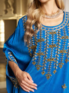 A woman wears the Social Queen Gilded Leaf Embellished Cape Dress in royal blue chiffon satin with gold and silver details, cape-style slit sleeves, and a layered necklace. She stands indoors with her face partially out of frame.