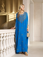 A woman wears the Social Queen Gilded Leaf Embellished Cape Dress in royal blue chiffon satin with gold leaf embroidery, cape-style slit sleeves, and pairs it with gold sandals and a small gold handbag by a white railing indoors.