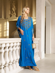 A woman stands indoors by a white balustrade, wearing the Social Queen Gilded Leaf Embellished Cape Dress in royal blue chiffon satin, with slit cape sleeves, holding a small handbag and looking to the side.