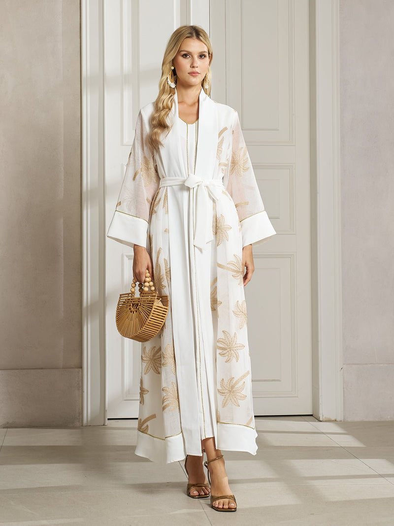 A woman indoors wears the Social Queen Gilded Palm Embroidered Sheer Jacket with beige floral patterns and a waist tie, paired with sandals and a wooden handbag—ideal for resort-inspired layering.