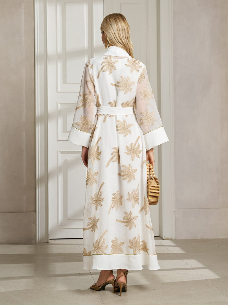 A woman stands indoors, facing away, wearing the Social Queen Gilded Palm Embroidered Sheer Jacket with beige palm leaf details and holding a wicker handbag.