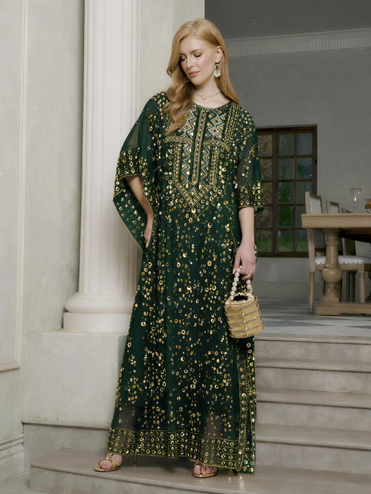 A woman indoors holds a woven handbag, wearing the Social Queen Gold Sequin Beaded Tulle Cape Dress. A set dining table and chairs are visible in the background.