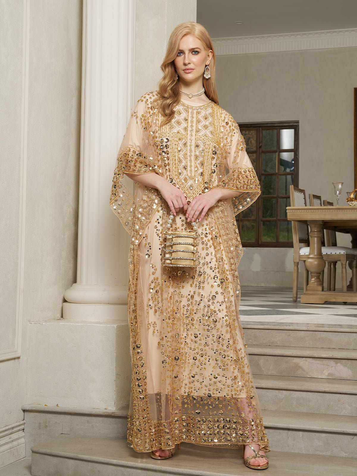 A woman stands on indoor steps wearing the Social Queen Gold Sequin Beaded Tulle Cape Dress and holding a matching purse, with a dining room in the background.