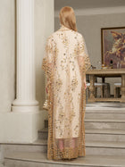 A woman with long blonde hair in the Gold Sequin Beaded Tulle Cape Dress by Social Queen stands on indoor steps, holding a small clutch, with a table and framed artwork visible in the background.