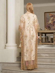 A woman with long blonde hair in the Gold Sequin Beaded Tulle Cape Dress by Social Queen stands on indoor steps, holding a small clutch, with a table and framed artwork visible in the background.