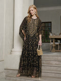 A woman stands on indoor stairs wearing the Social Queen Gold Sequin Beaded Tulle Cape Dress, gold earrings, sandals, and holding a small woven handbag.