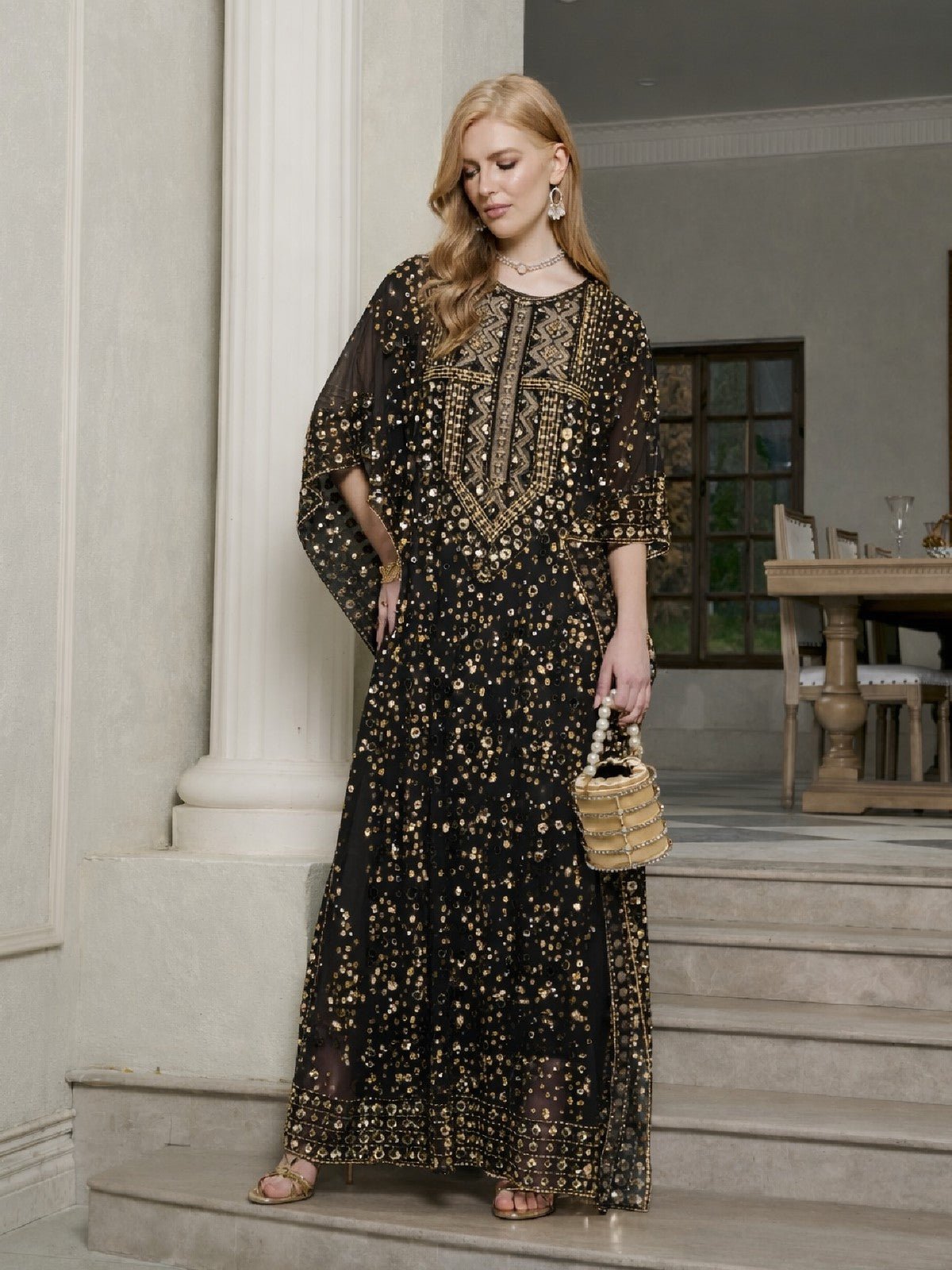 A woman stands on indoor stairs wearing the Social Queen Gold Sequin Beaded Tulle Cape Dress, gold earrings, sandals, and holding a small woven handbag.