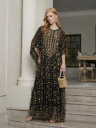 A woman stands on indoor stairs wearing the Social Queen Gold Sequin Beaded Tulle Cape Dress, gold earrings, sandals, and holding a small woven handbag.