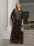 A woman stands on indoor steps in the Social Queen Gold Sequin Beaded Tulle Cape Dress, accented with a beaded handbag, earrings, and sandals—an elegant and unique choice for wedding guests.