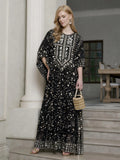 A woman stands on indoor steps wearing the Social Queen Gold Sequin Beaded Tulle Cape Dress, paired with earrings, sandals, and a small handbag.