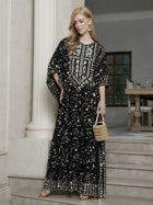 A woman stands on indoor steps wearing the Social Queen Gold Sequin Beaded Tulle Cape Dress, paired with earrings, sandals, and a small handbag.