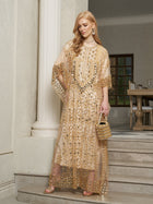A woman wearing the Social Queen Gold Sequin Beaded Tulle Cape Dress stands on marble steps indoors, accessorizing with a small gold bag and statement earrings.