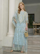A woman in a Social Queen Gold Sequin Beaded Tulle Cape Dress stands on marble stairs, holding a gold handbag, with a dining room in the background.