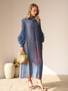 A woman indoors holds a wooden handbag and wears gold heels with the Social Queen Gradient Pleated Dress with 3D Floral Sleeves. Two ceramic vases with plants are behind her—an ideal, stylish look for a wedding guest.