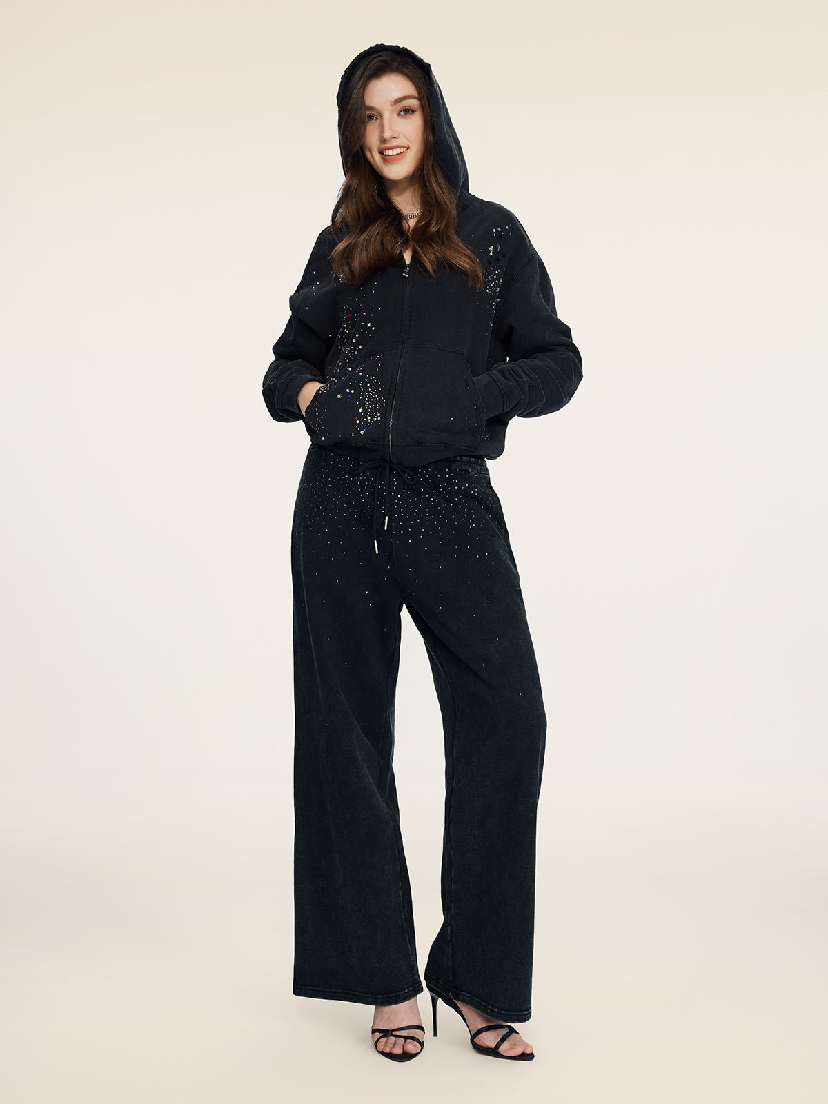 A person stands facing forward in Social Queen's Gradient Rhinestone Wide-Leg Pants with an elastic waist, a black hooded zip-up jacket, and black high-heeled sandals.