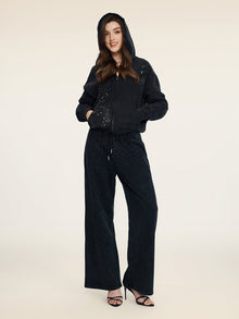 A person stands facing forward in Social Queen's Gradient Rhinestone Wide-Leg Pants with an elastic waist, a black hooded zip-up jacket, and black high-heeled sandals.